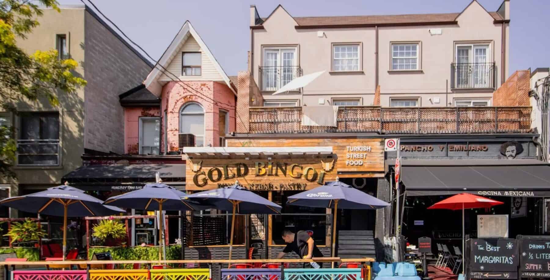 Vibrant street scene in Kensington Market with outdoor seating, colorful storefronts, and a sign for Gold Bingo, showcasing Toronto's trendy neighbourhood atmosphere.