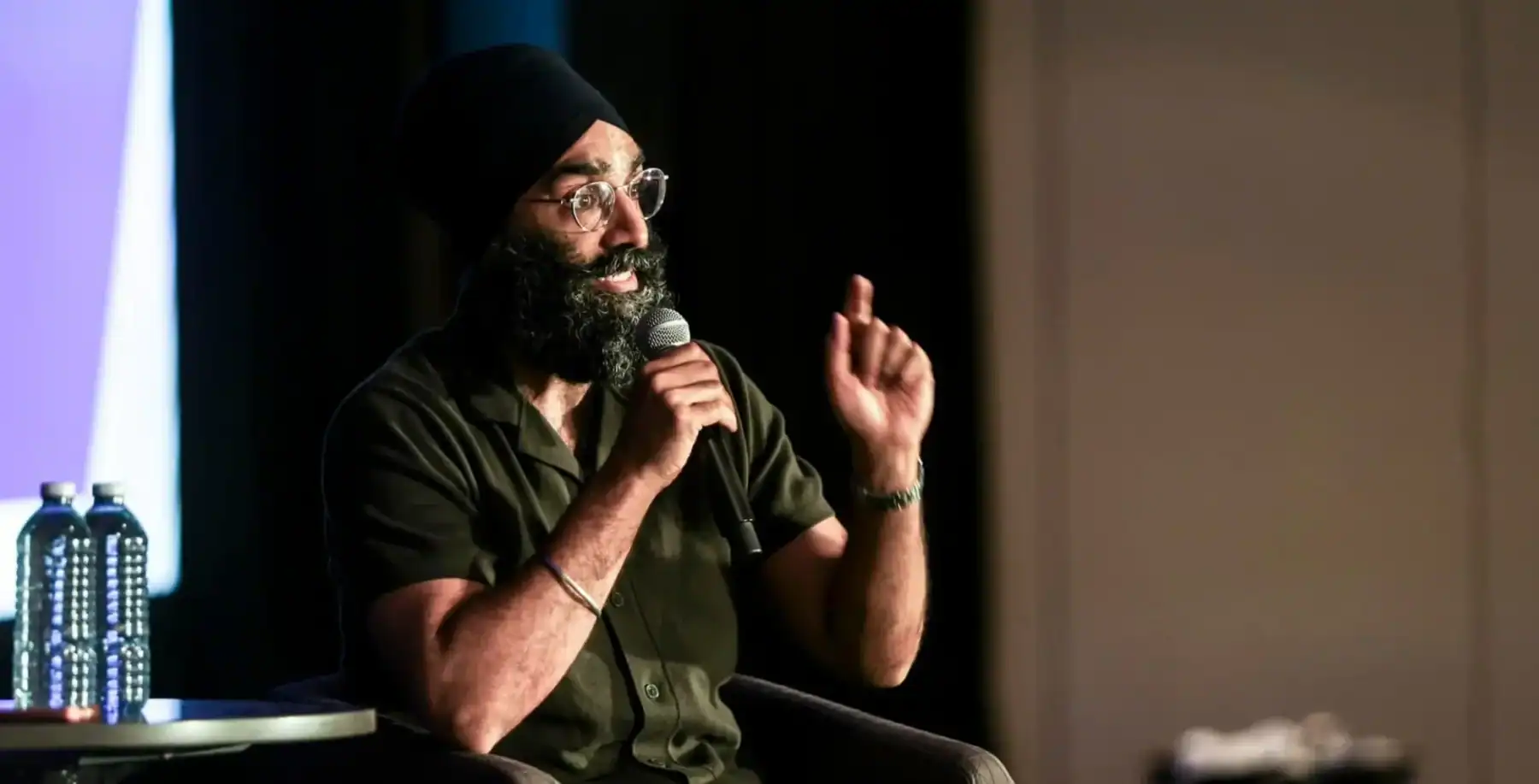 Jagmeet Singh's lawyer brother speaking at an event, wearing a turban and glasses, holding a microphone, with a focused expression.