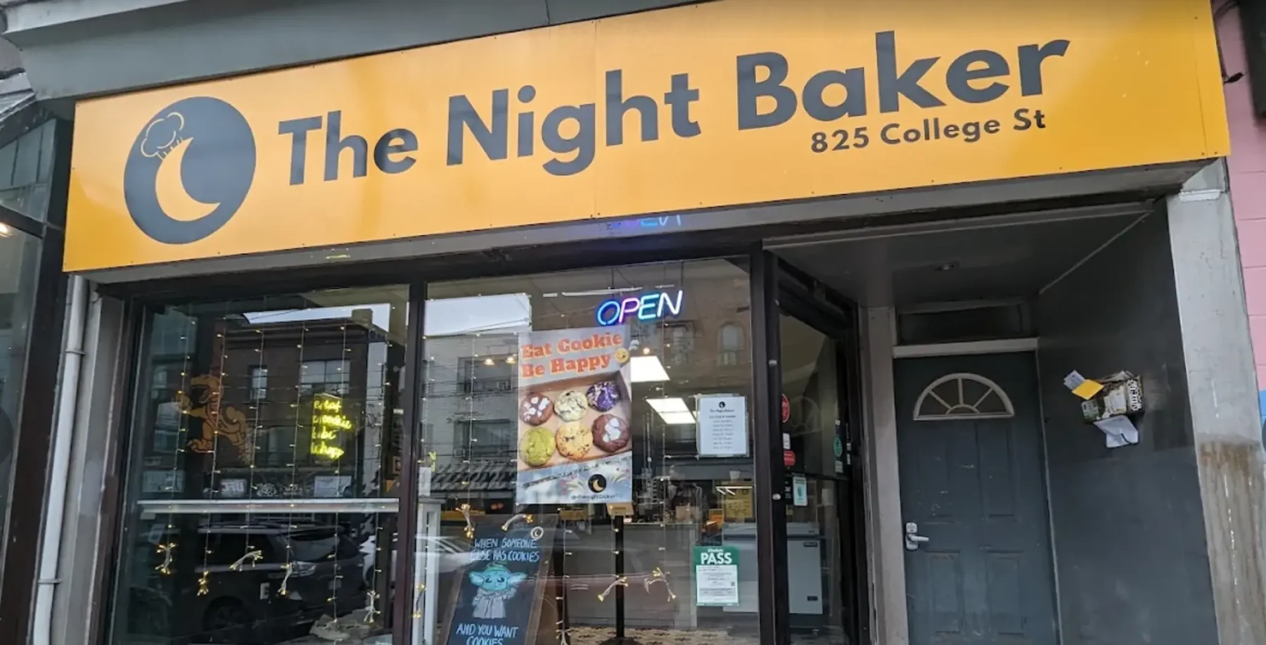 The Night Baker bakery storefront on College Street with a yellow sign and glass door, featuring a poster and open sign, in Toronto.