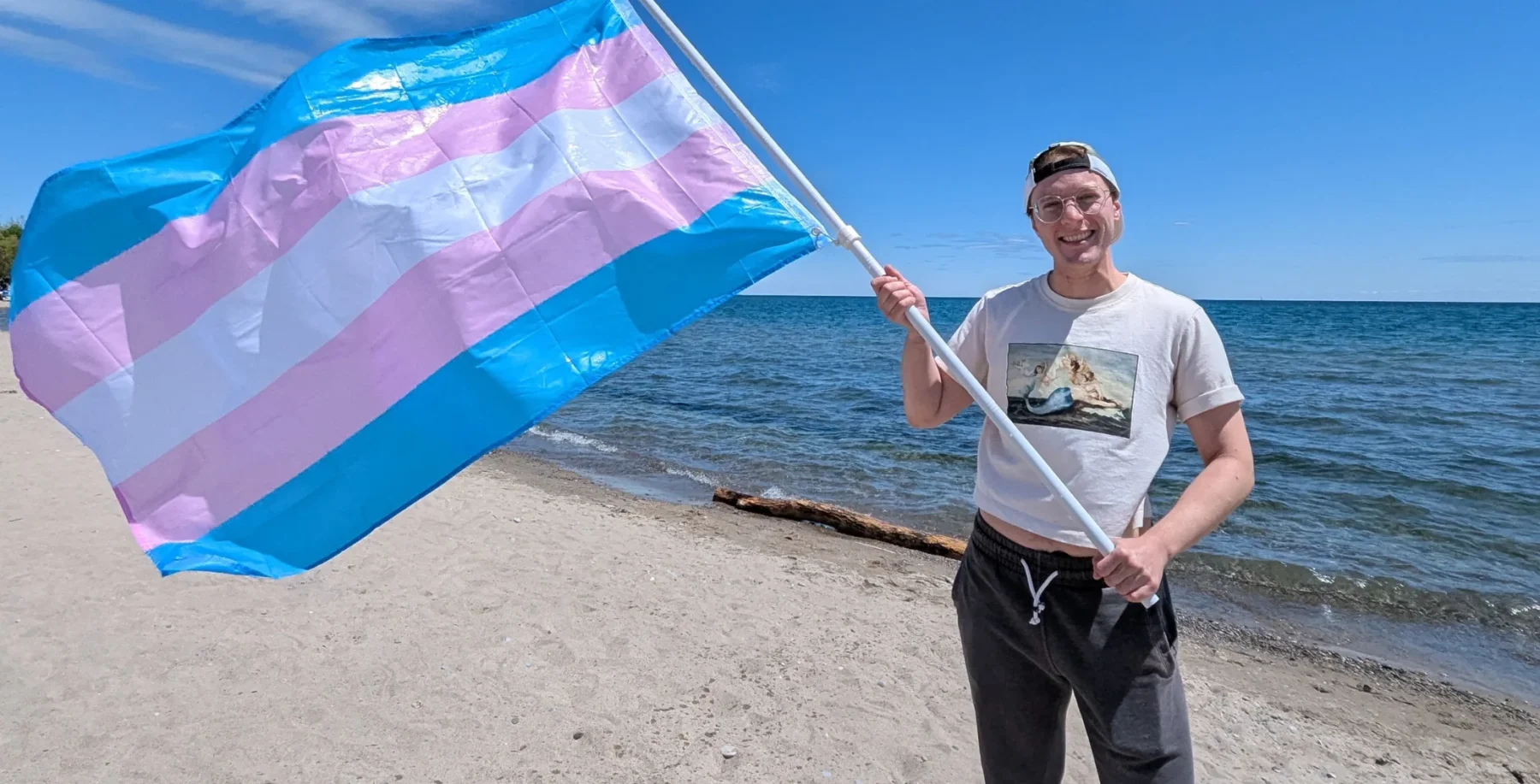 Transgender Toronto athlete holding a large transgender pride flag on a sandy beach with the lake and blue sky in the background.