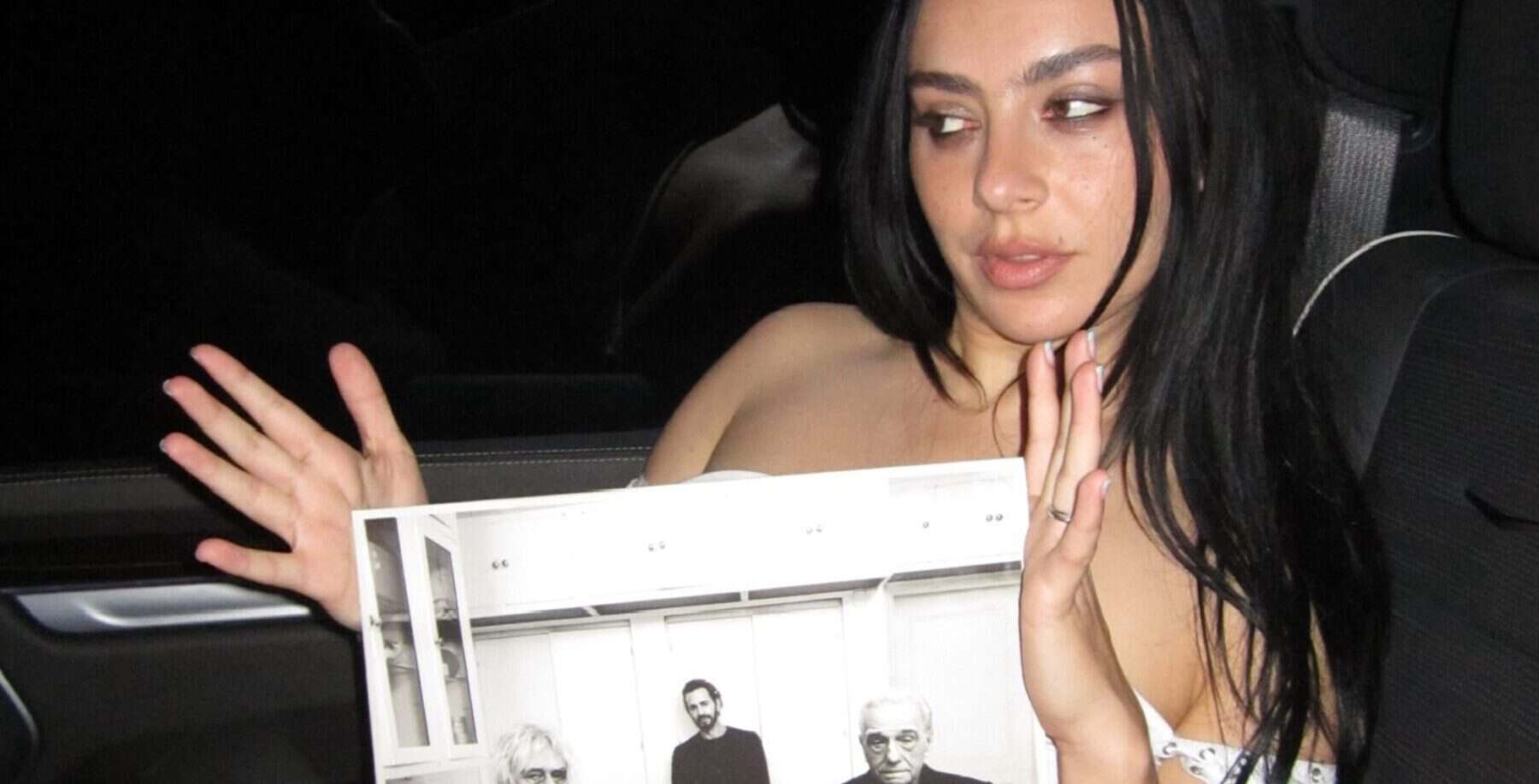 Young woman with dark hair in a car holding a black-and-white photo, looking to the side with a thoughtful expression.