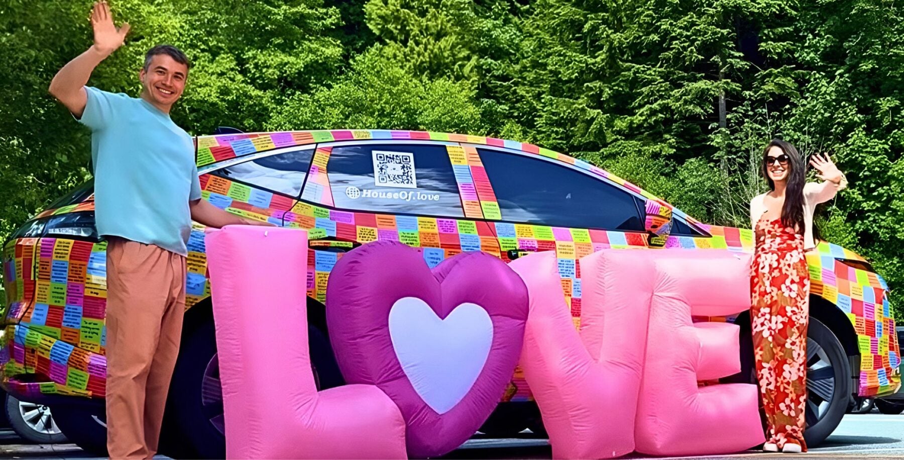 Two people standing next to a colorful car with a large inflatable 'LOVE' sign in front, surrounded by green trees, celebrating love and community in Toronto.