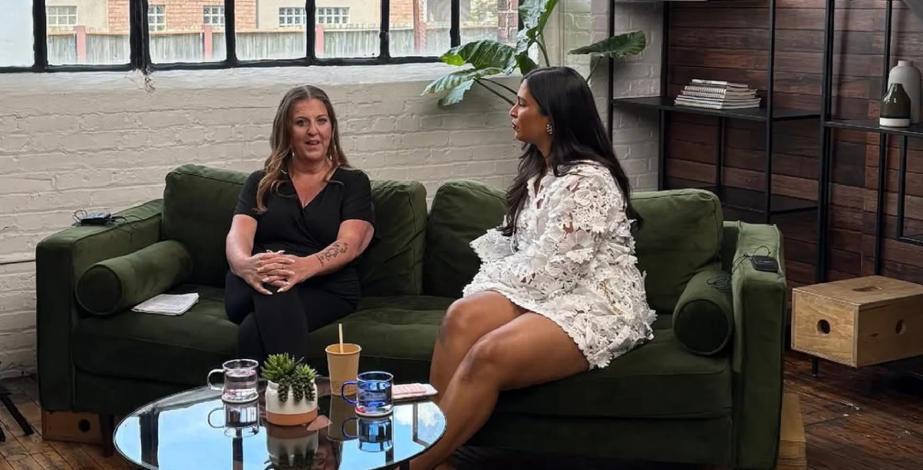 Two women having a conversation on a green sofa in a modern, well-lit studio with large windows and indoor plants.