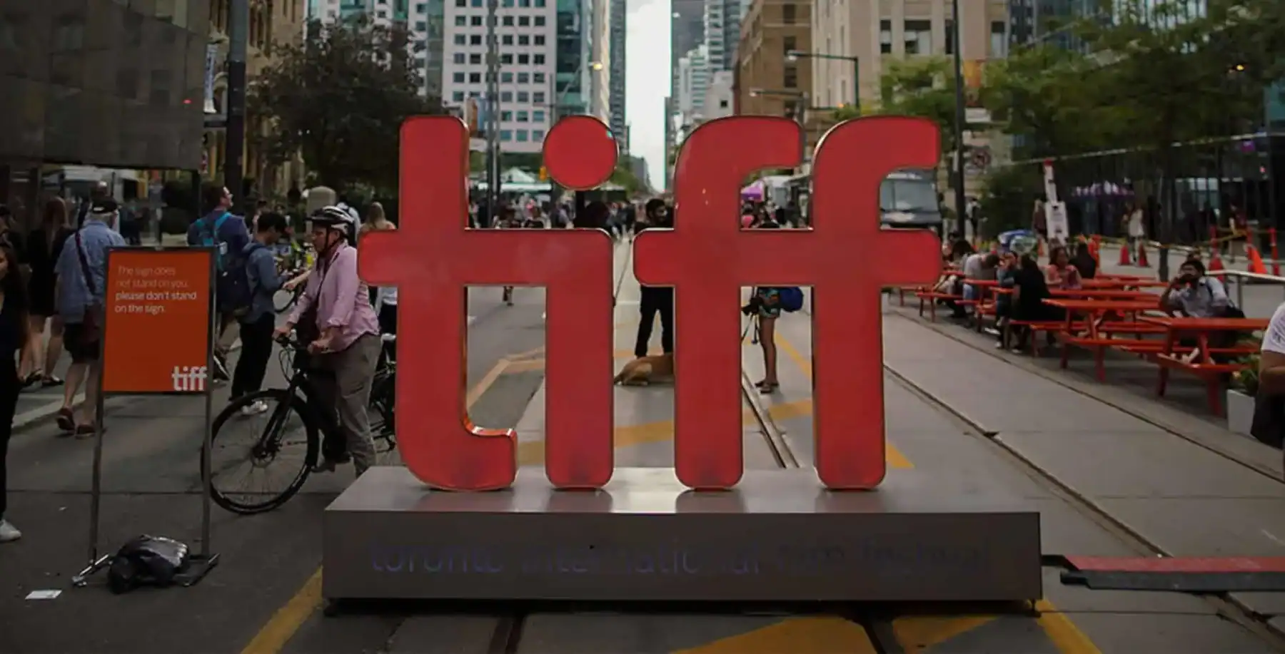 Toronto street scene with large red 'tiff' sign in the foreground, surrounded by pedestrians, cyclists, and city buildings, representing the Toronto International Film Festival.