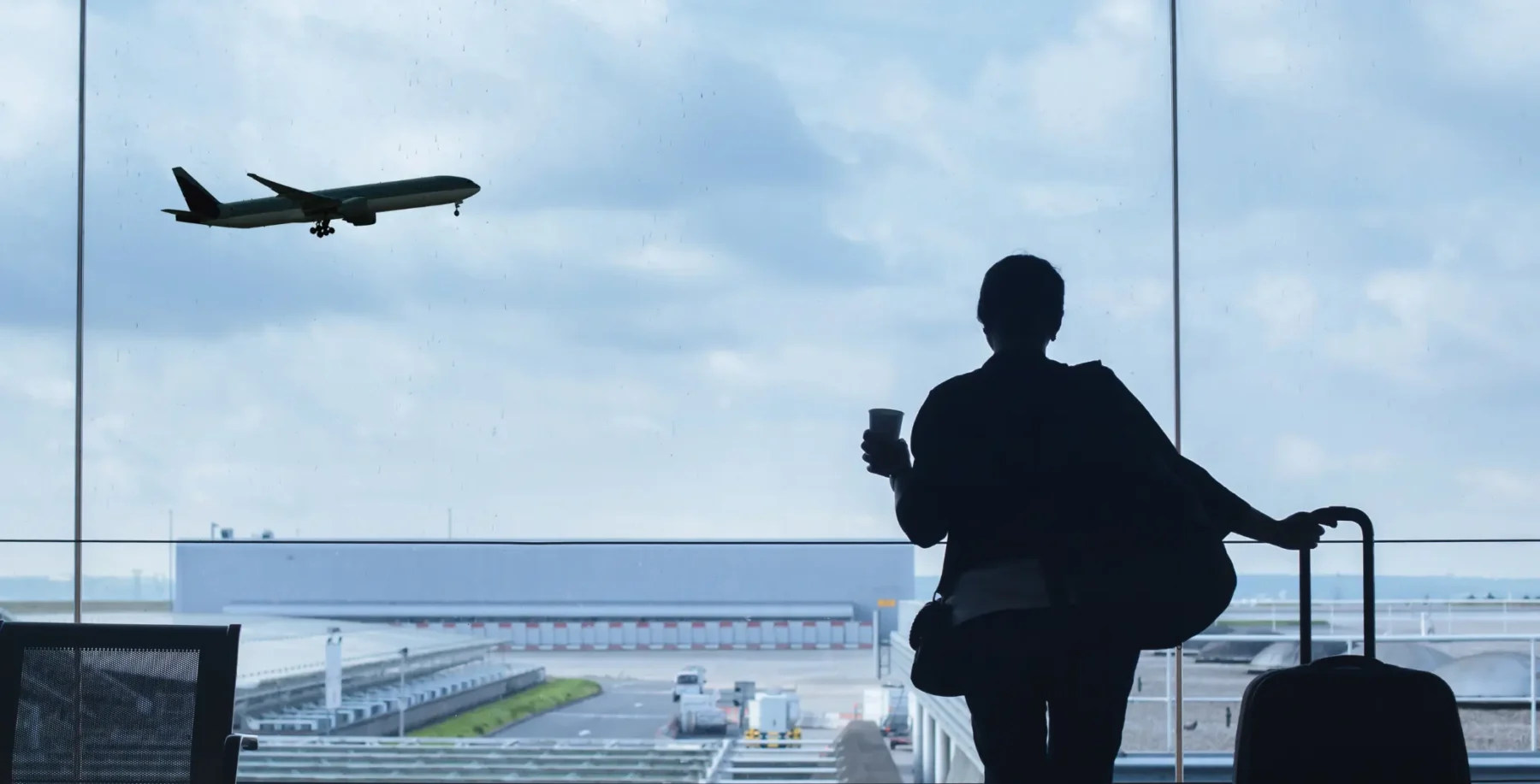 Silhouette of a traveler with a suitcase and a drink at an airport terminal, with an airplane taking off in the background.