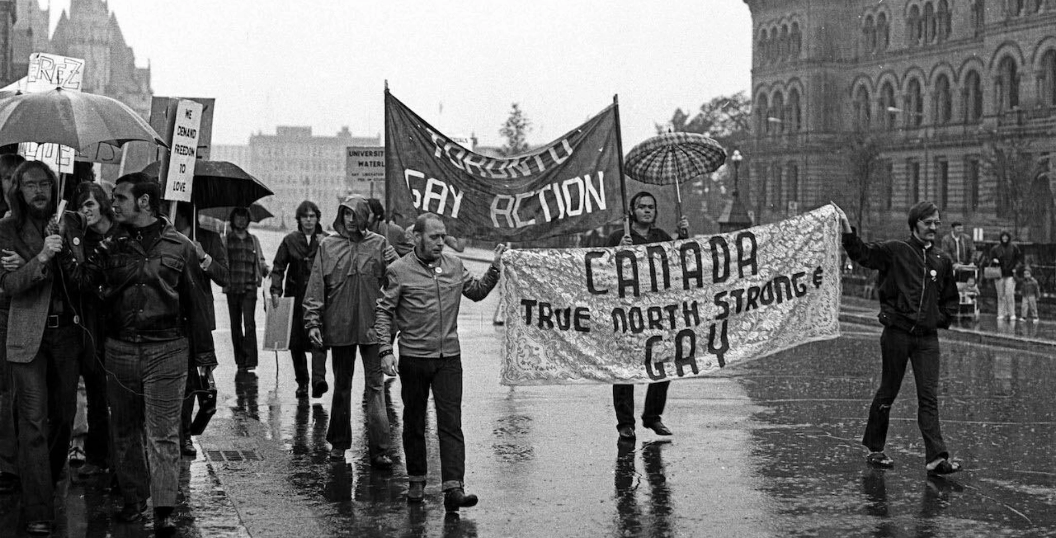 LGBTQ+ pride protesters march in the rain holding banners, including one that reads 'Canada True North Strong Gay,' during a Pride protest event.