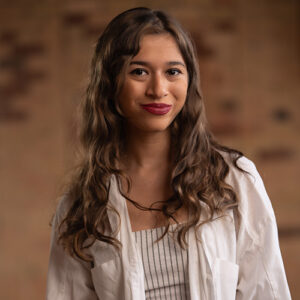 Young woman with long wavy brown hair wearing a white blazer and striped top, smiling softly against a warm, blurred background, representing Toronto lifestyle and culture.