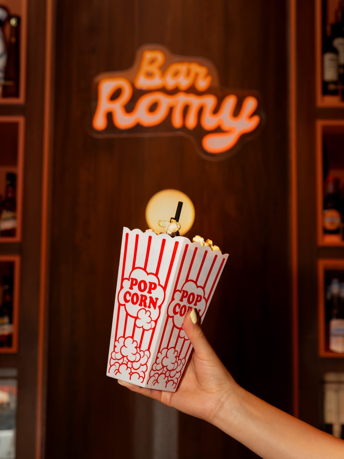 Hand holding a popcorn bucket in front of a bar sign at TIFF event, with warm lighting and wooden decor.