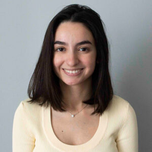 Young woman with shoulder-length dark hair smiling at the camera, wearing a cream-colored top, against a plain light gray background.