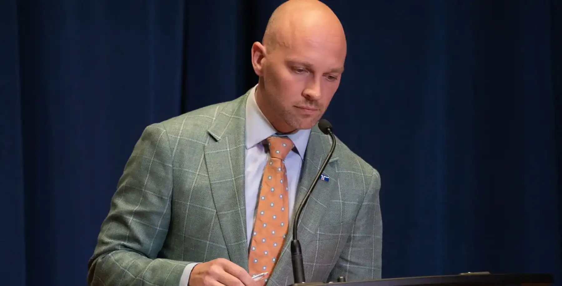 Toronto mayoral candidate Brad Bradford speaking at a podium during a campaign event, wearing a grey checkered suit and orange tie.