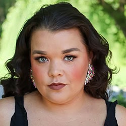 Close-up portrait of a young woman with dark wavy hair, makeup, and earrings, outdoors with green foliage in the background, representing diverse Toronto community.
