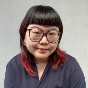 Close-up of a young woman with black and red hair, wearing glasses and a dark top, smiling softly against a plain background, representing diverse Toronto community.