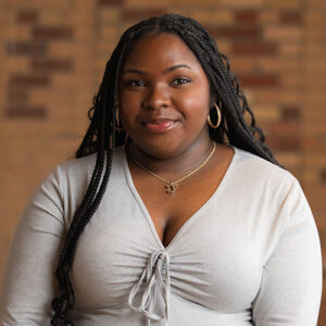 Portrait of a confident Black woman with long braided hair, wearing a light grey top and gold jewelry, standing against a blurred brick wall background.