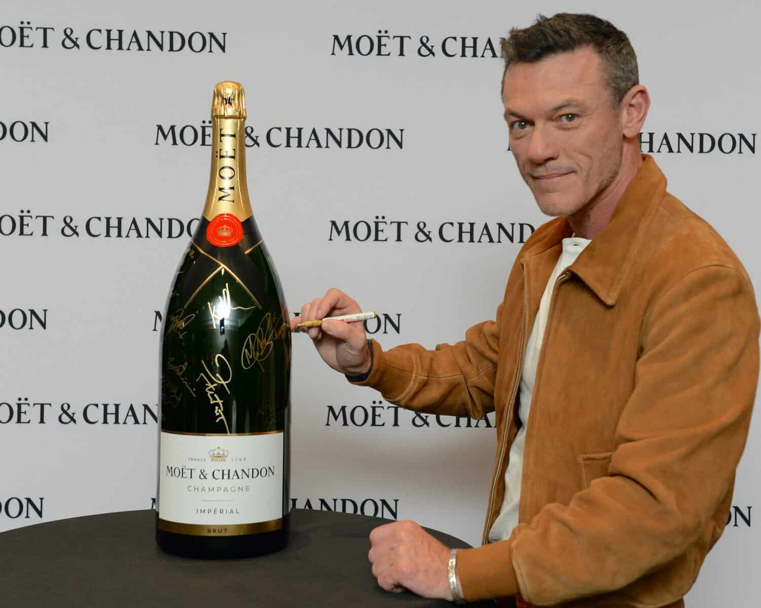 Man signing a large Moët & Chandon champagne bottle at TIFF event with branded backdrop in the background.