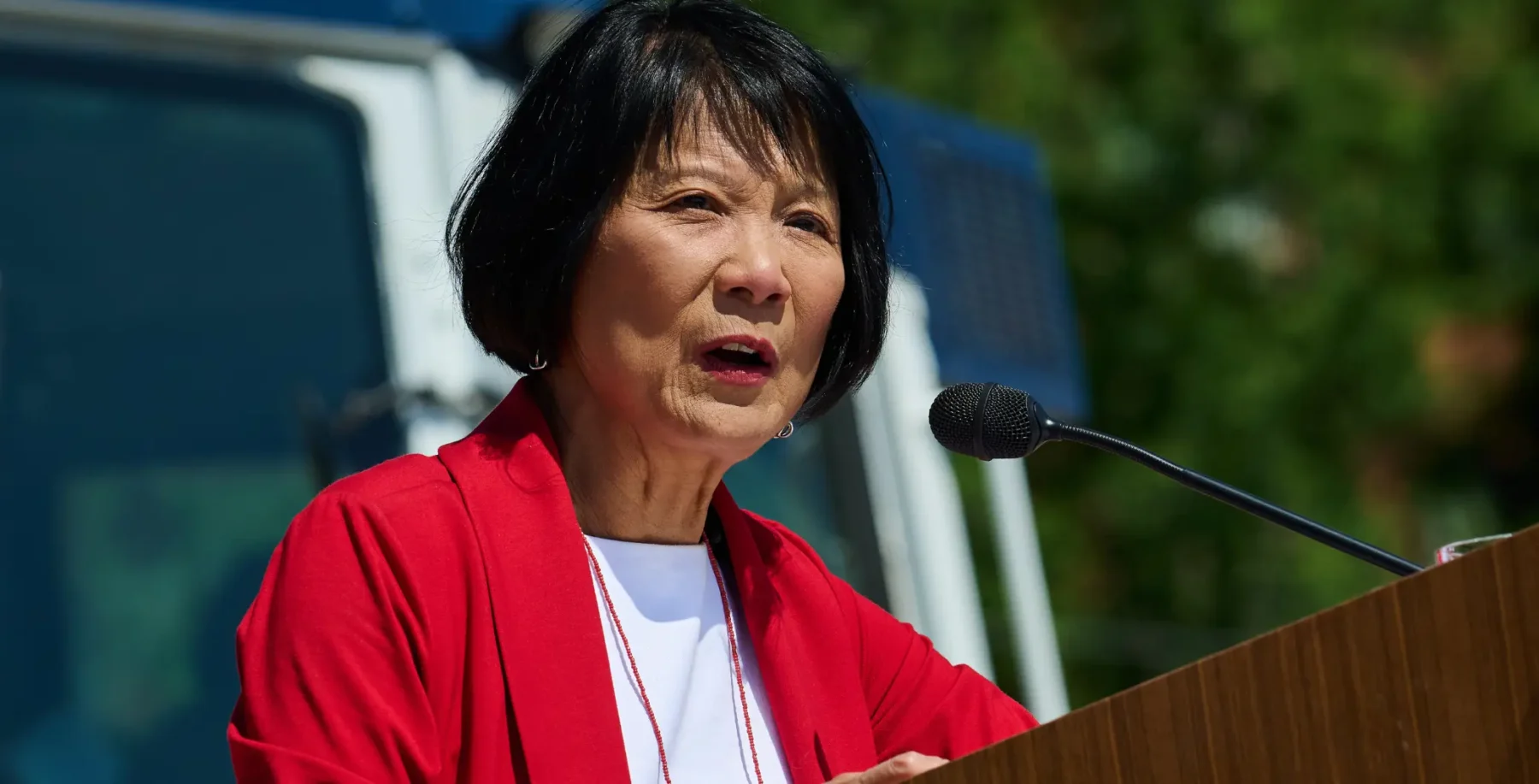Asian woman speaking at a podium outdoors, wearing a red jacket and white shirt, with a microphone in front of her, against a background of trees and a vehicle.