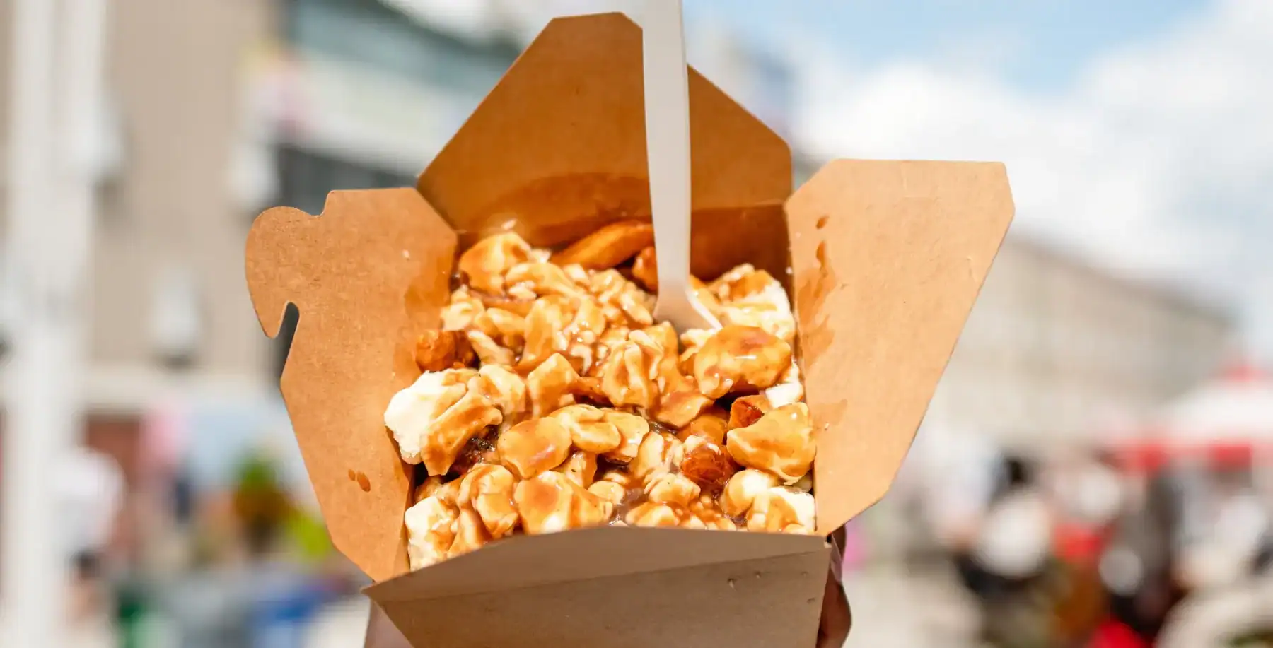 Close-up of a cardboard container filled with caramel popcorn, held outdoors during daytime with a blurred city street background.