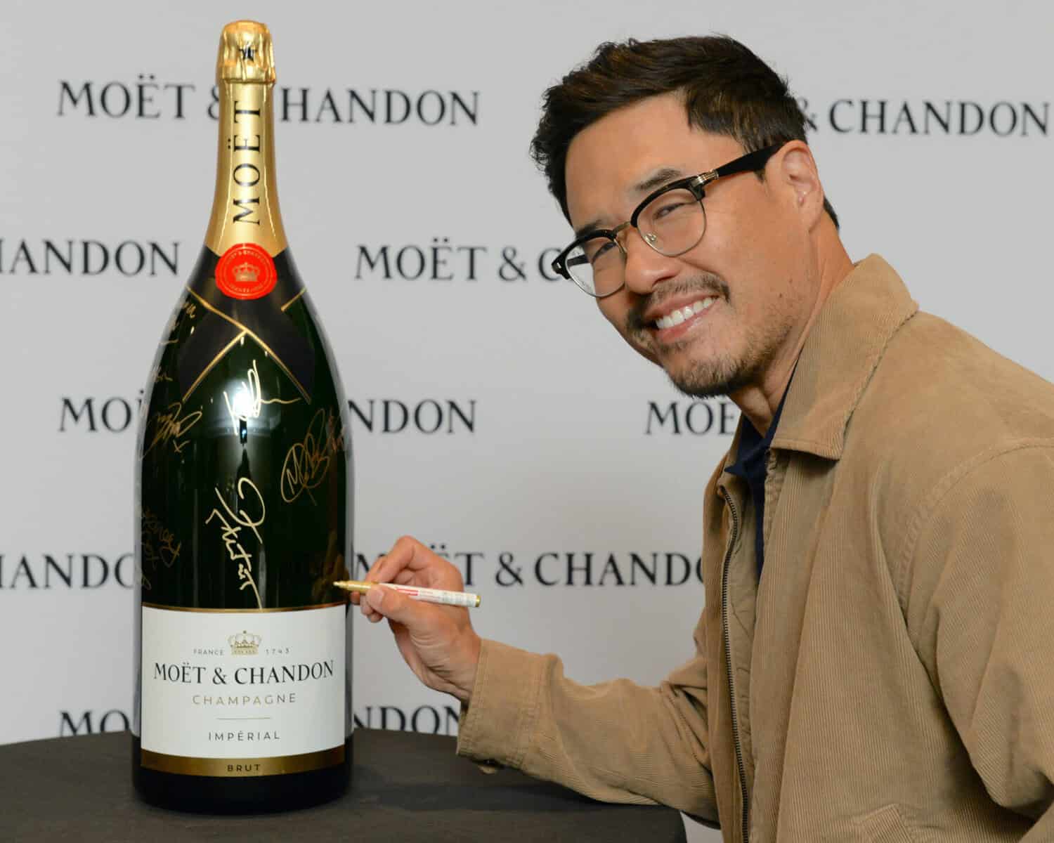 Man signing a large Moët & Chandon champagne bottle at TIFF event, smiling, wearing glasses and a tan jacket.