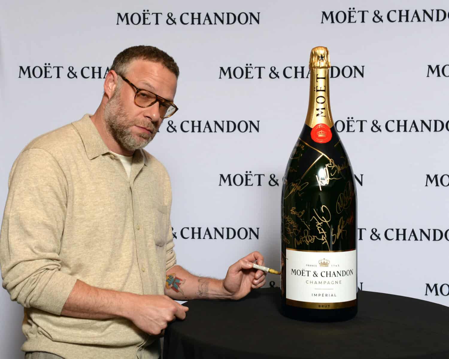 Man with glasses and beard signing a large Moët & Chandon champagne bottle at TIFF event.