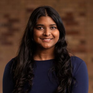 Smiling woman with long dark hair wearing a navy blue top against a blurred brick wall background, representing Toronto's diverse community.