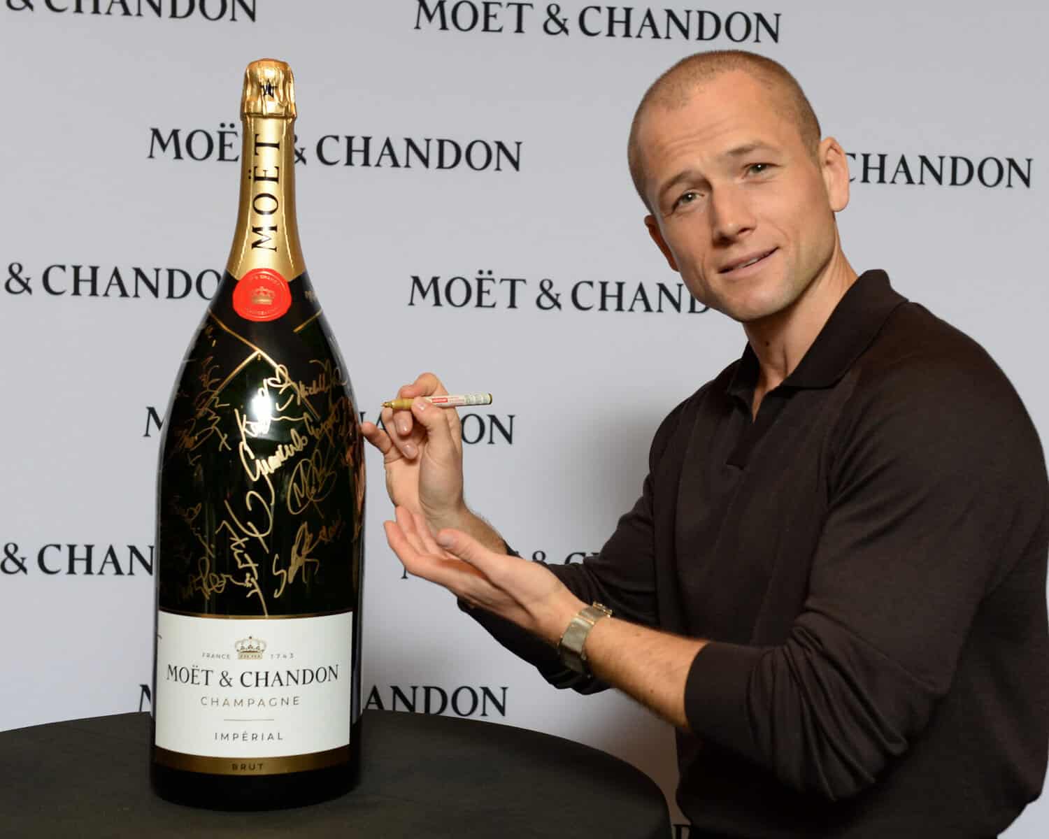 Man signing a large Moët & Chandon champagne bottle at TIFF event with a branded backdrop in the background.