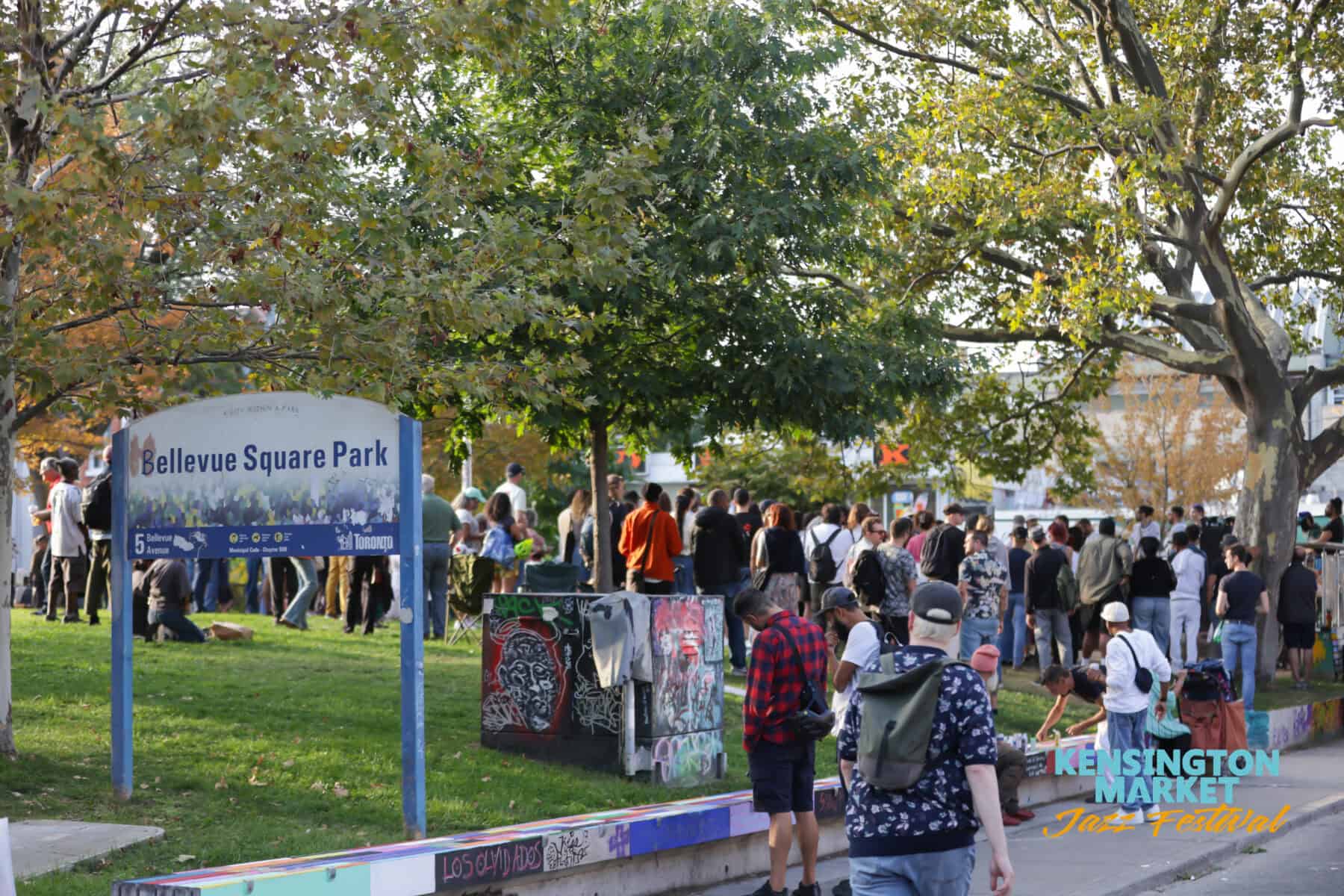 Crowd gathered at Bellevue Square Park during the Kensington Market Jazz Festival in Toronto, with trees and festival signage visible.