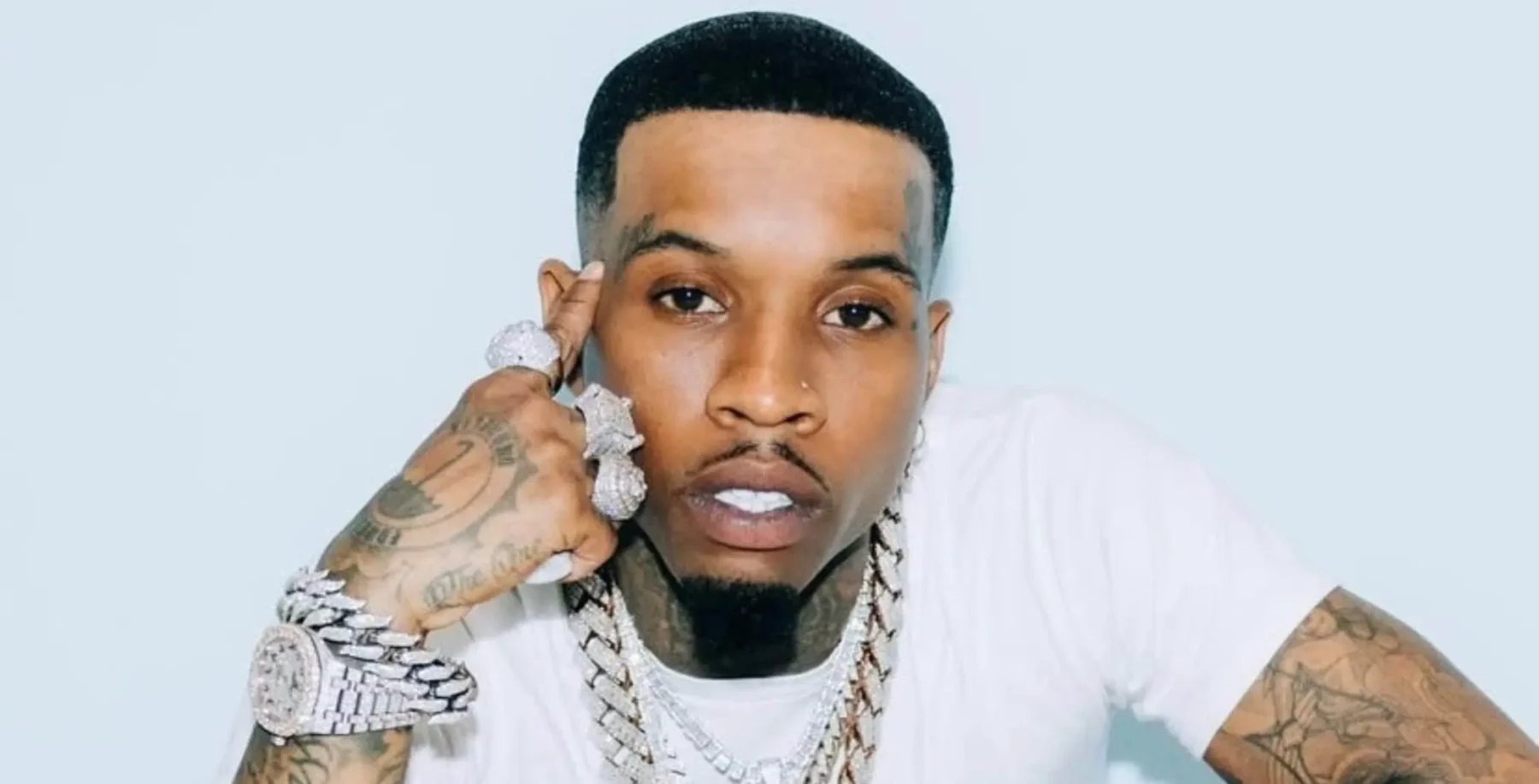 Close-up of a young man with tattoos and jewelry, wearing a white t-shirt, looking directly at the camera against a light blue background.