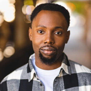 Portrait of a young Black man with short curly hair and a beard, wearing a plaid shirt, outdoors in Toronto with blurred city lights in the background.