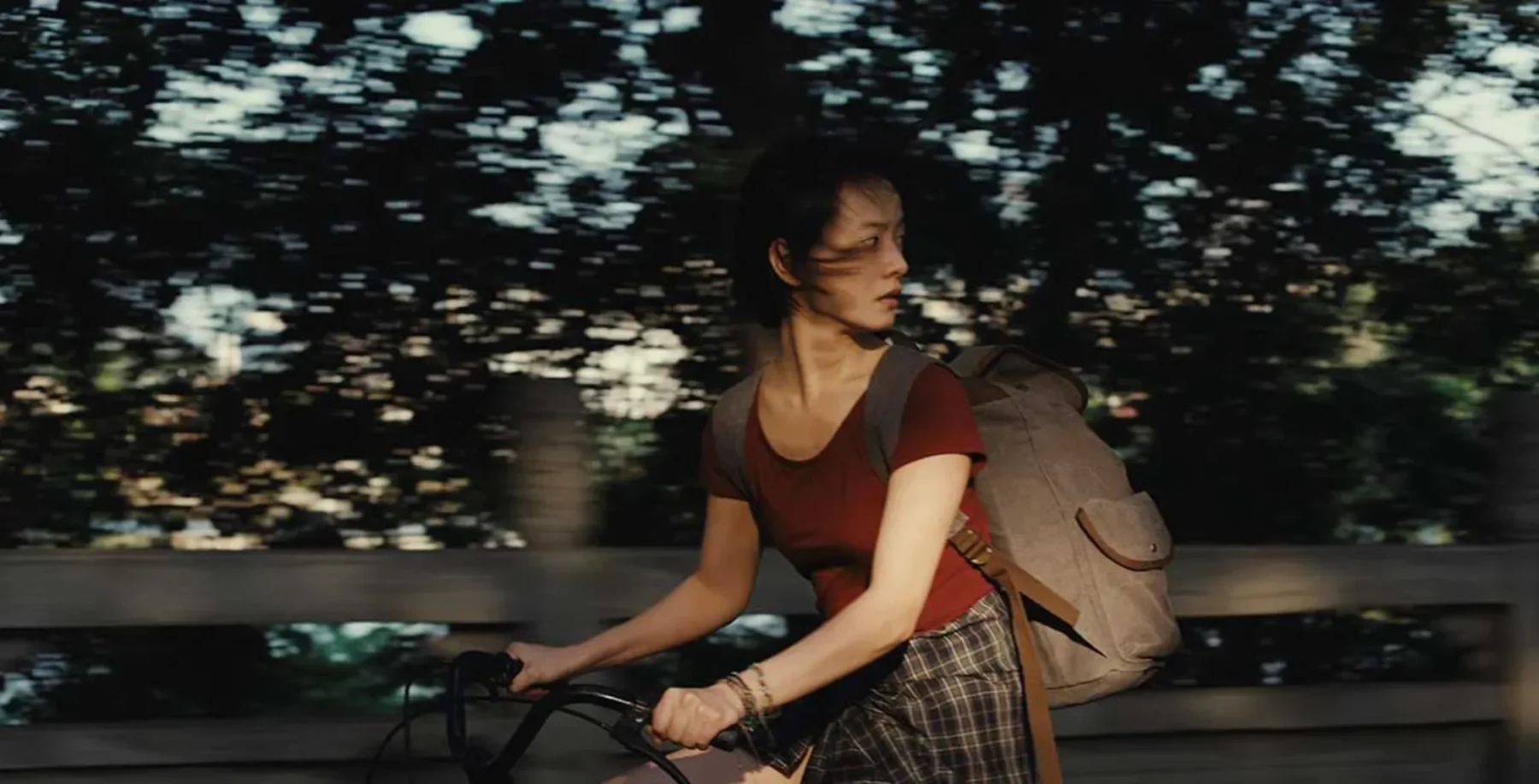 A young woman with dark hair riding a bicycle on a city street at dusk, carrying a backpack, with blurred trees and streetlights in the background.