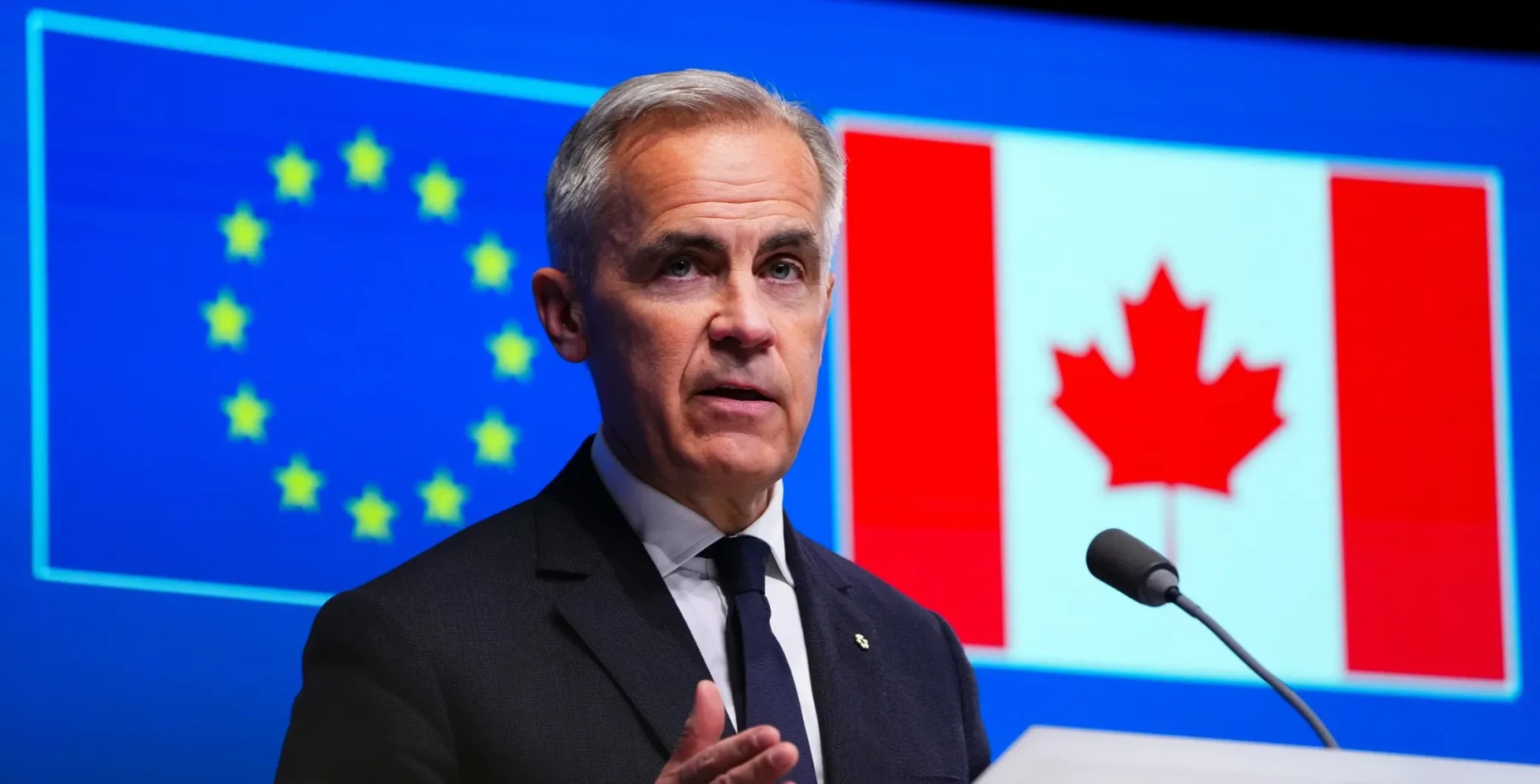European Union flag and Canadian flag displayed behind a man speaking at a podium during a press conference.