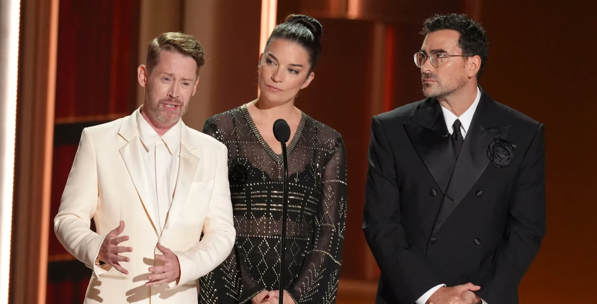 Three individuals standing at a podium during an awards ceremony, with a warm wooden background, dressed in formal attire, engaging in a discussion.