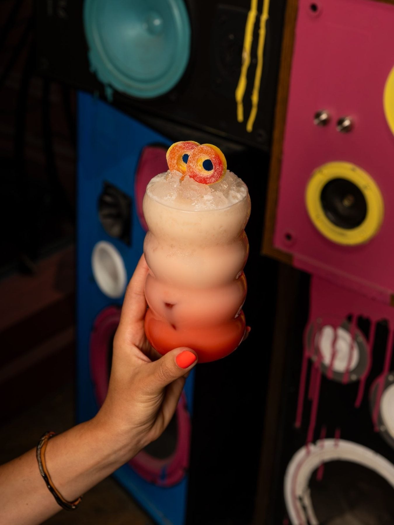 Colorful cocktail with a gummy worm garnish held in front of a vibrant, retro-style speaker setup at TIFF event.