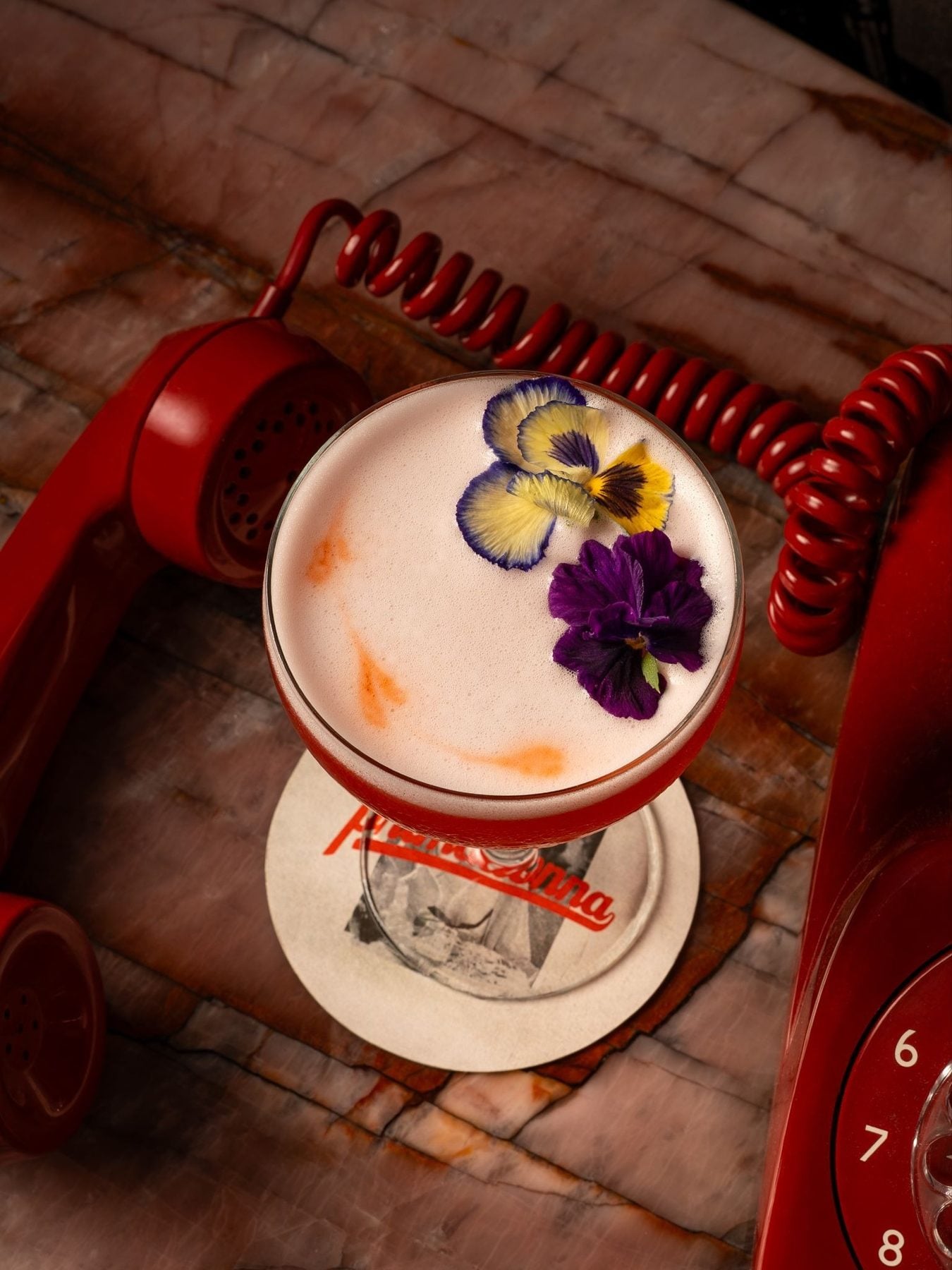 A cocktail garnished with edible flowers, served in a glass on a wooden surface with vintage red telephone and clock, inspired by TIFF screenings.