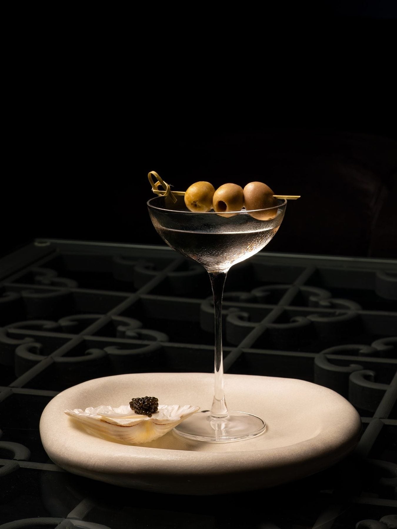 Elegant cocktail glass with green olives on a skewer, served on a white plate against a dark background, highlighting a sophisticated drink presentation.