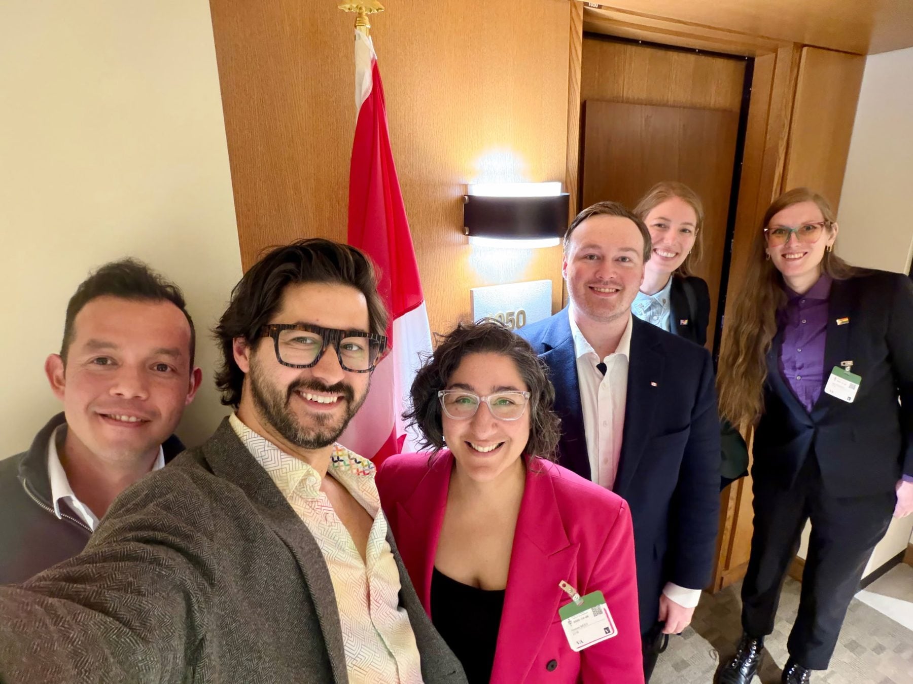 Group of diverse 2SLGBTQIA+ activists and allies gathering on Parliament Hill for a rally demanding $450M federal investment, with Canadian flag in background.