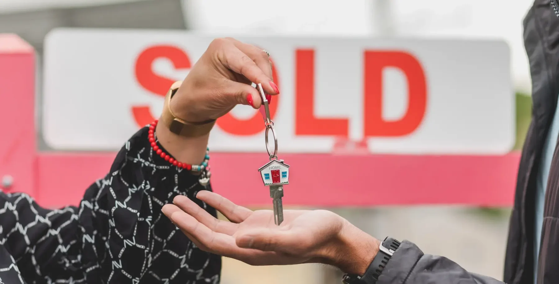 Person handing over a house key to another person in front of a 'SOLD' sign, symbolizing real estate transaction and housing market activity.