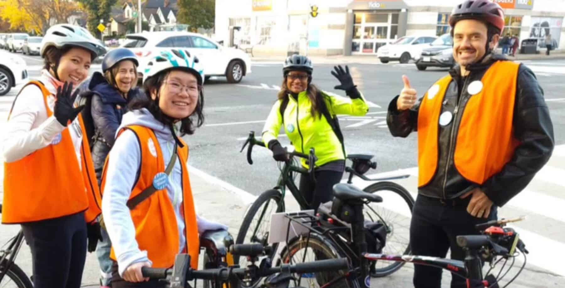 Cyclists in orange safety vests wave at the camera, ready to ride to polling stations as part of Bike Bus event in Toronto.