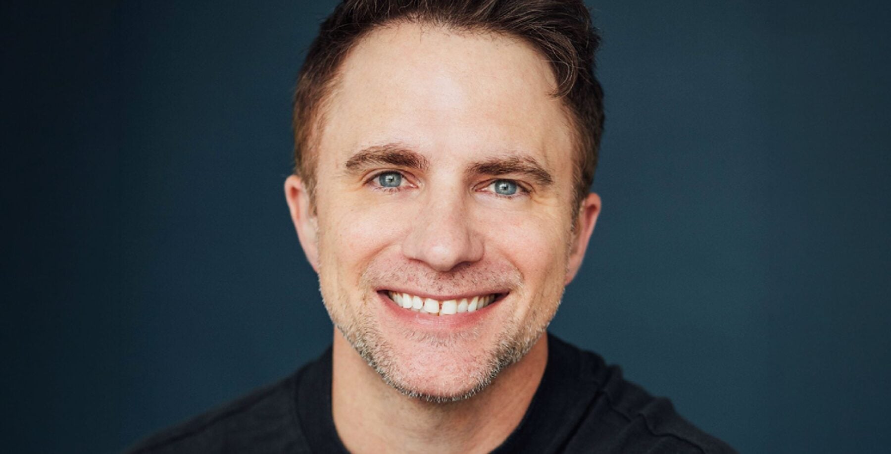 Close-up of D.J. Demers smiling, wearing a black shirt against a dark background, at Just For Laughs Toronto comedy event.