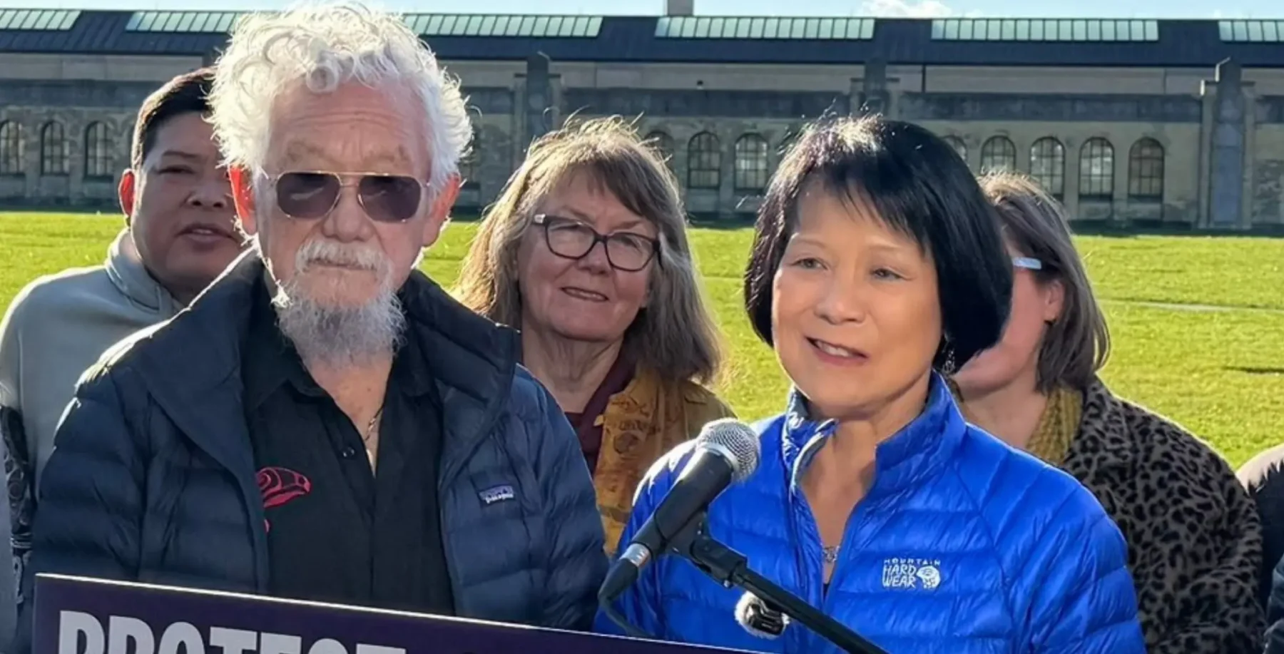 A diverse group of people, including a woman speaking at a microphone outdoors, with a historic building in the background, during a public event in Toronto.