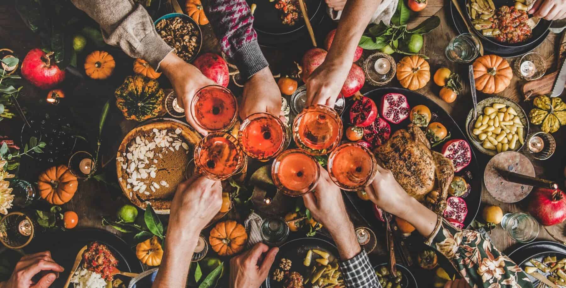 People clinking glasses of drinks at a festive Friendsgiving feast with pumpkins, fall decorations, and a variety of food on the table.