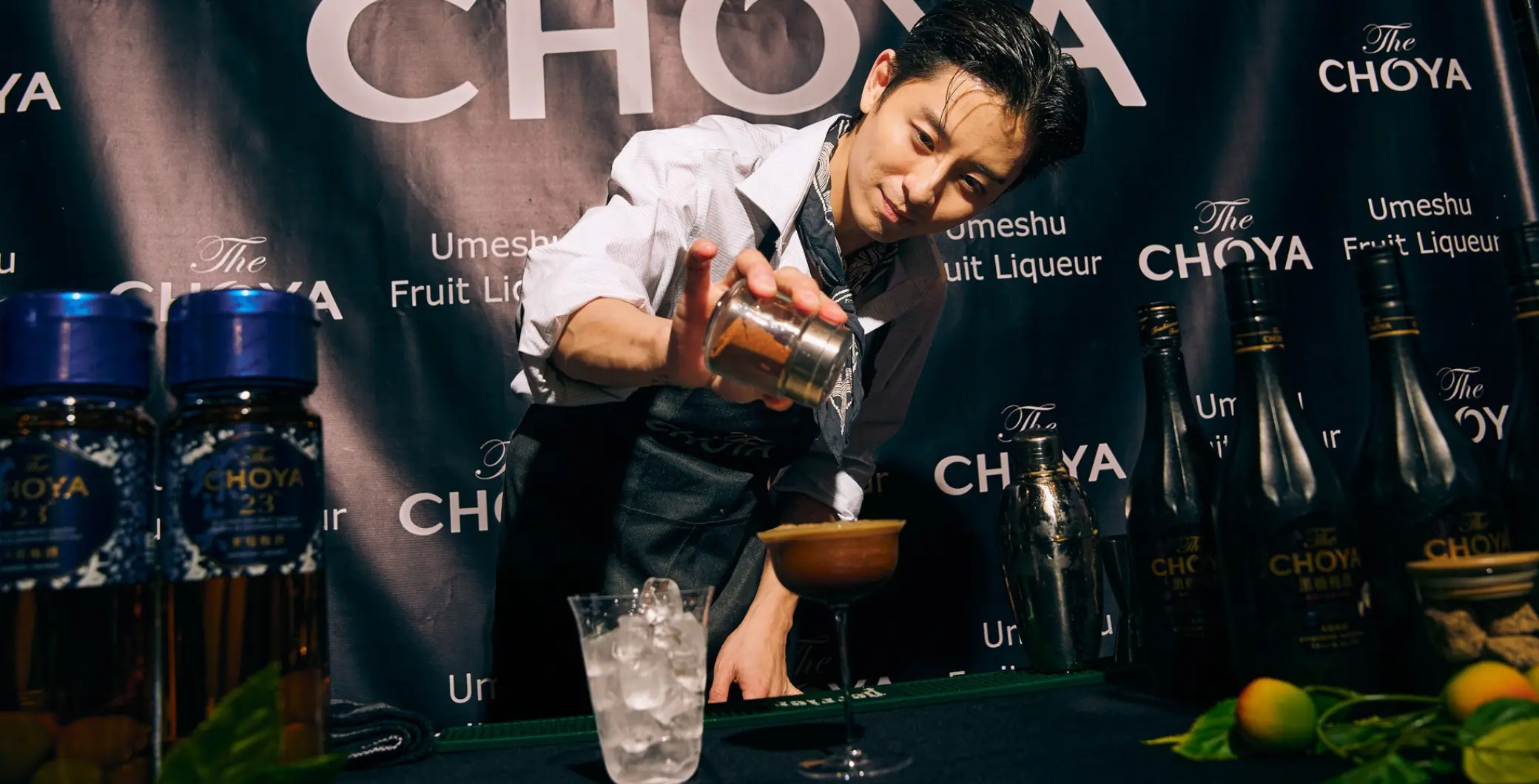 Barman pours a cocktail at a branded NOW Toronto event setup with The Choya bottles and lime garnish visible in the background.