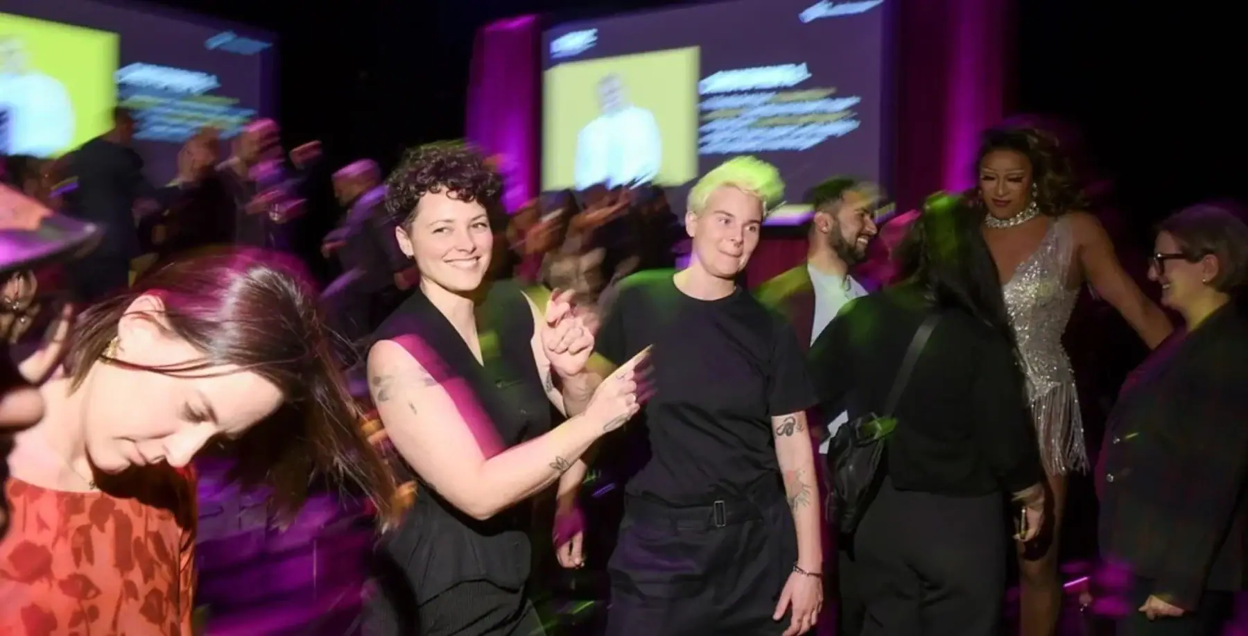 Group of diverse people dancing and enjoying a lively event with colorful lighting and screens in the background at NOW Toronto.