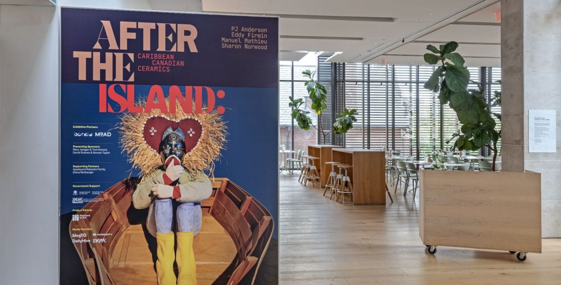 NOW Toronto exhibit poster 'After the Island' displayed near a wooden reception cart in a bright gallery space with plants and large windows.