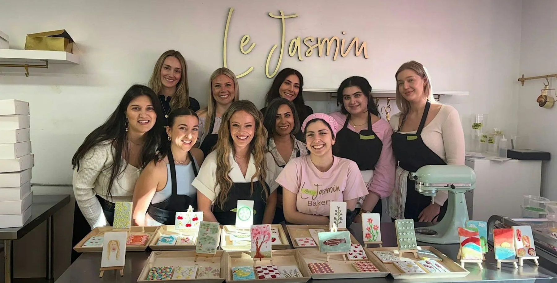 Group of women in a bakery workshop, smiling behind a table with decorated baked goods, in a bakery named Le Jasmin, celebrating a baking class in Toronto.