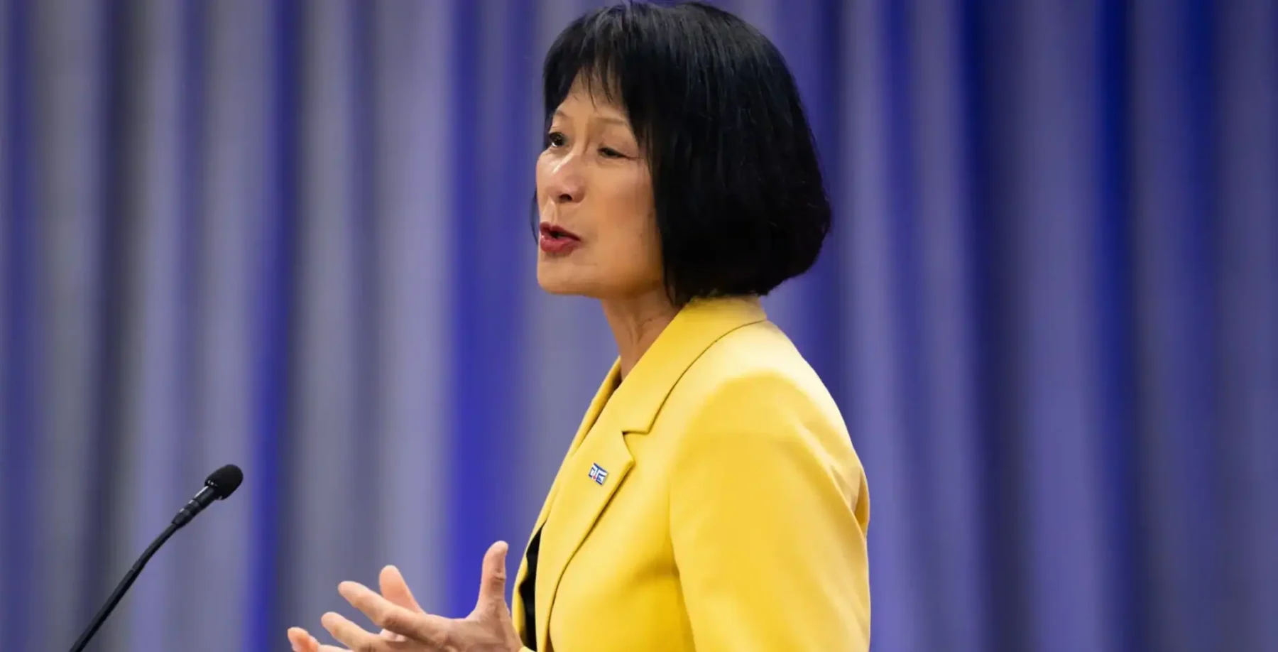Olivia Chow speaking at a podium in a bright yellow blazer during a press event in Toronto, addressing the audience with a focused expression.