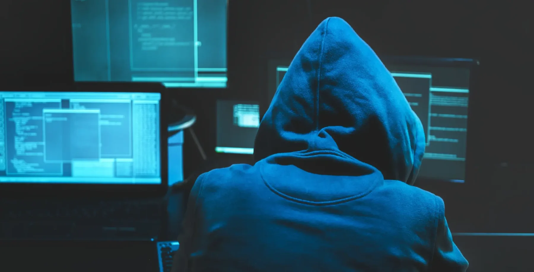 Hacker in a hoodie working on multiple computer screens with code, highlighting cybersecurity threats and digital security concerns.