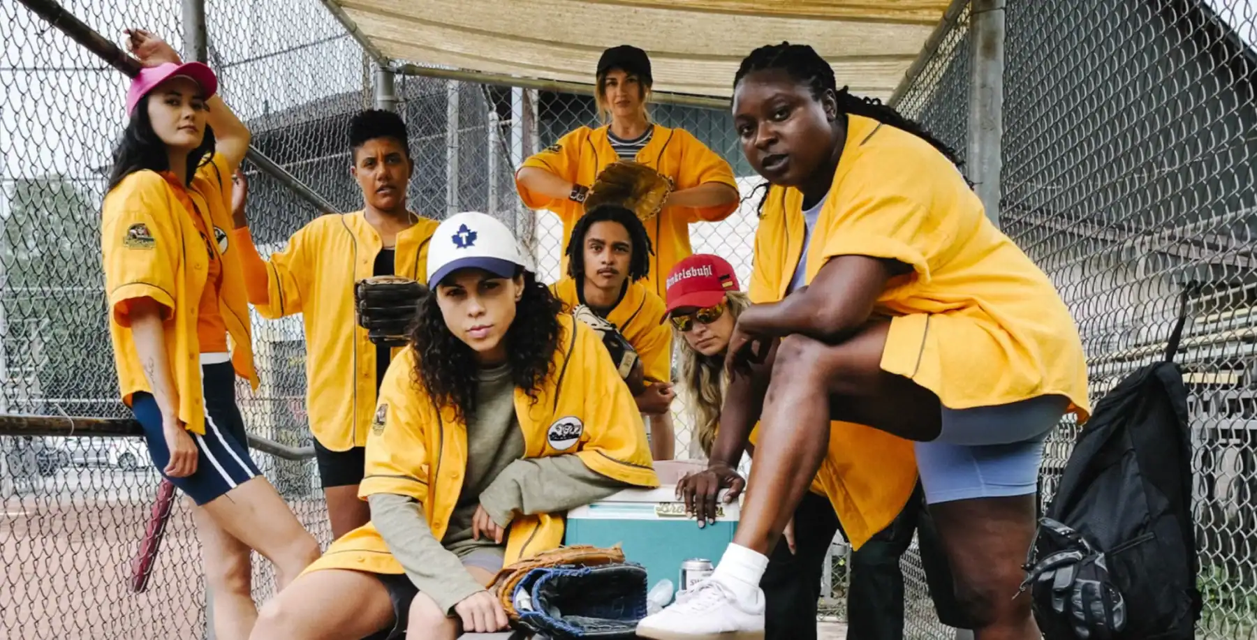 Diverse group of young women and men in yellow softball uniforms on a baseball field, preparing for a game, with a chain-link fence in the background.