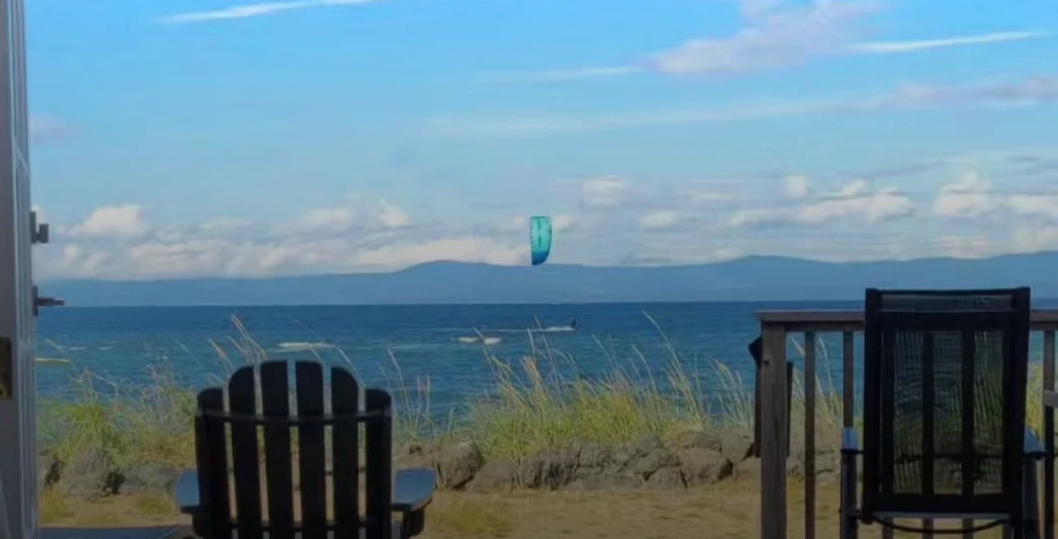 Scenic view of the ocean with a kite in the sky, two chairs facing the water, and grassy shoreline, capturing a peaceful coastal landscape in Vancouver Island.