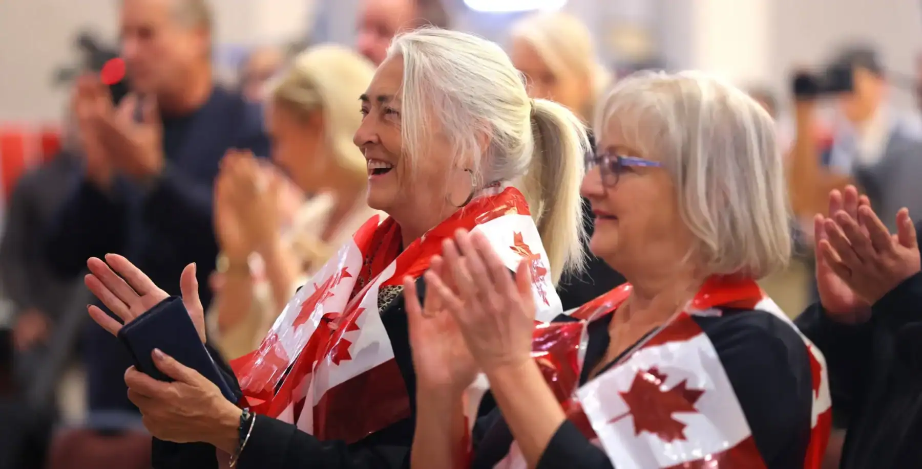 Two elderly women with Canadian flags enjoying a public event, smiling and clapping, representing Canadian patriotism and community spirit.