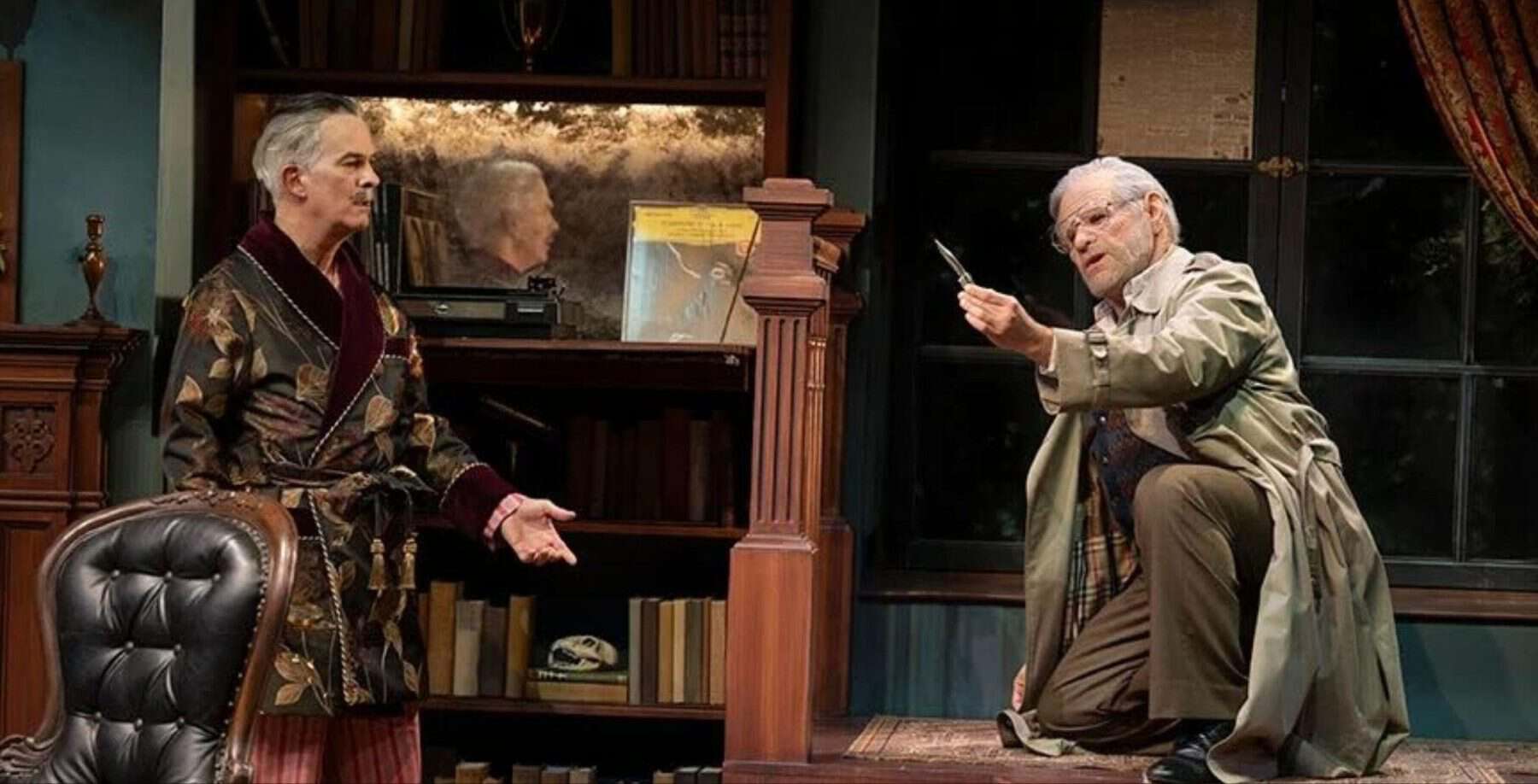 Two actors performing a scene on stage in a vintage-style setting with bookshelves and a window, capturing a dramatic moment in a theatrical play.