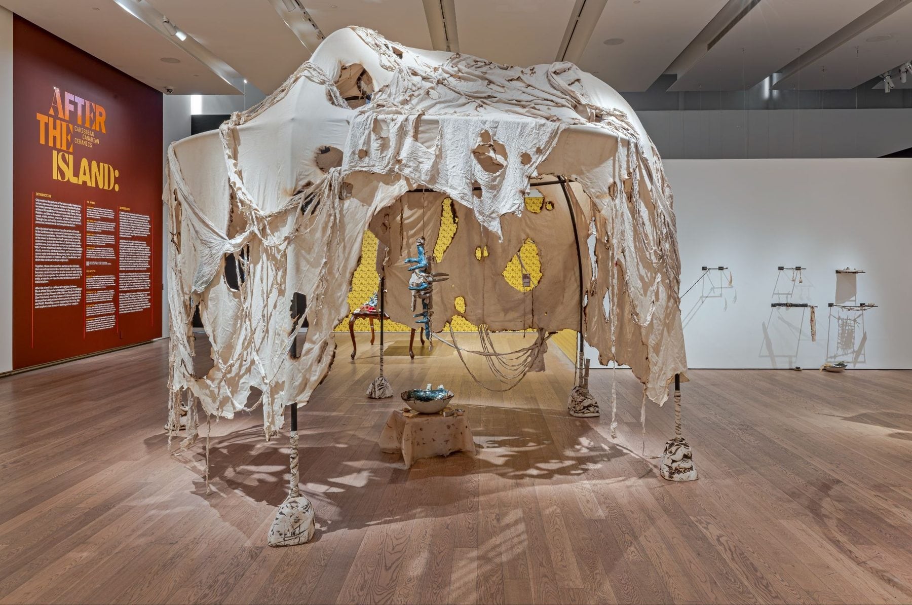 NOW Toronto exhibit at Gardiner Museum featuring a sculptural installation with tattered fabric, suspended framework, and scattered objects on a wooden gallery floor in a modern exhibit space.